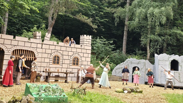 Menschen in mittelalterlichen Kostümen versammeln sich vor einer Burg, die im Wald liegt.