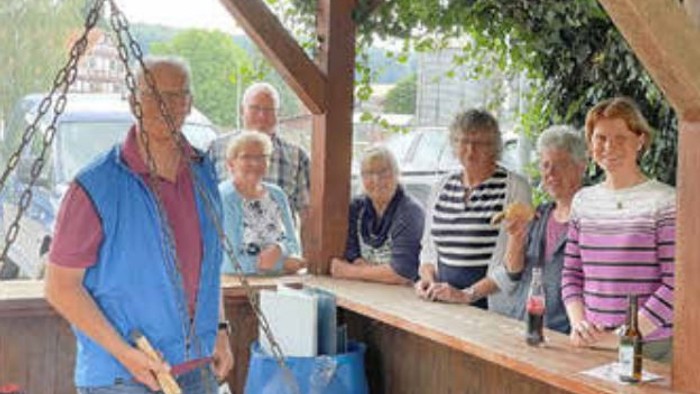 Gruppe von Menschen, die an einer Holzbartheke versammelt sind, einige halten Getränke, im Vordergrund ein Mann in einer blauen Weste.