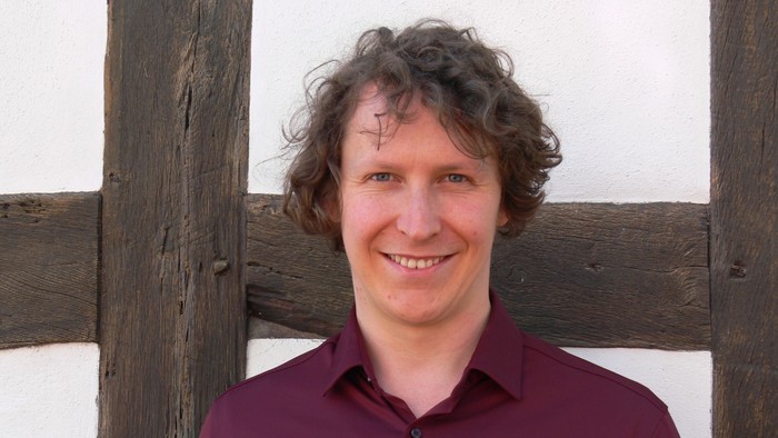 Mann im bordeauxroten Hemd steht vor hölzernen Balken.