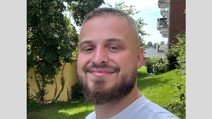 En ung mand med kort hår og skæg står i en have med græs og buske. I baggrunden er der et gult hus og et rødt boligkompleks.