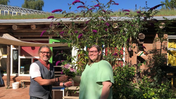 Zwei Menschen lächeln vor einem Gartenhaus mit Blumen.