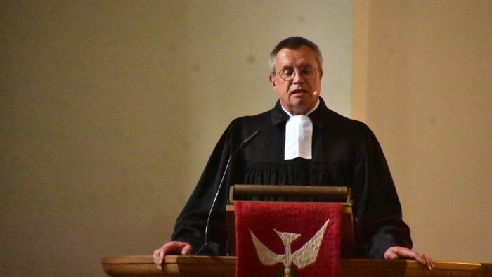 Ein Priester steht hinter einem Redepult mit Kreuzsymbol.