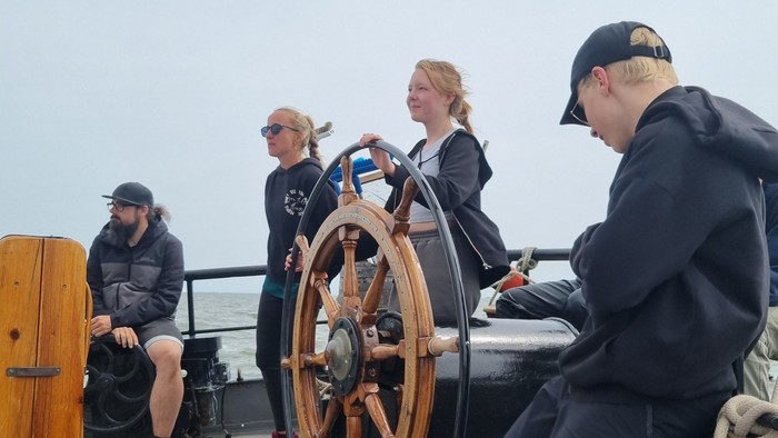 Vier Personen auf einem Boot, eine lenkt mit einem großen hölzernen Steuerrad.