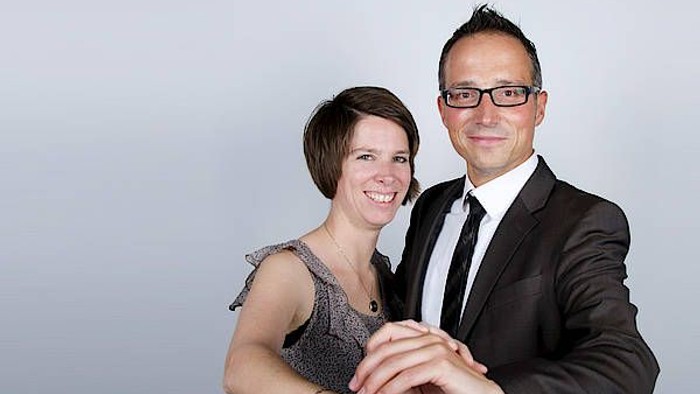 Ein Mann und eine Frau posieren für ein Foto, er trägt Anzug und Krawatte, sie ein graues Kleid.