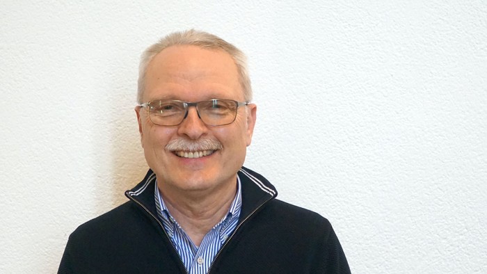 Ein älterer Mann mit Brille und schwarzer Jacke lächelt in die Kamera.