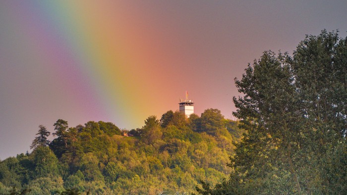 Ein Turm auf einem Hügel, umgeben von Bäumen bei Sonnenuntergang mit einem Regenbogen am Himmel.