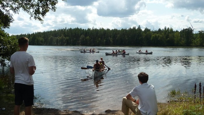 Zwei Männer sitzen am Seeufer und beobachten Ruderer auf dem Wasser.