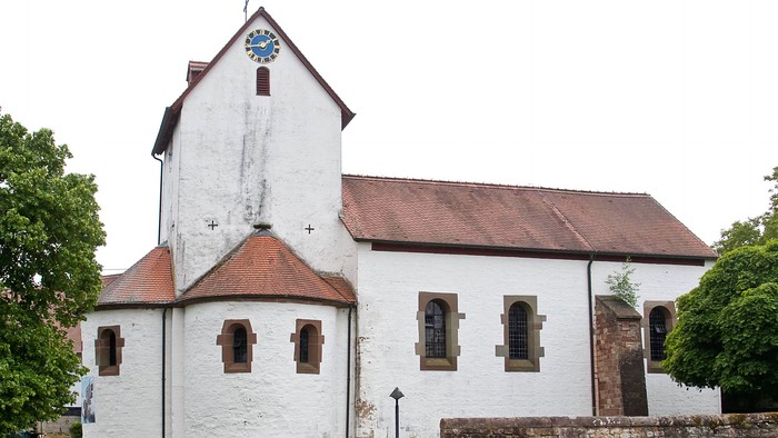 Historische weiße Kirche mit Uhrturm, umgeben von Grünflächen und einer Steinmauer.