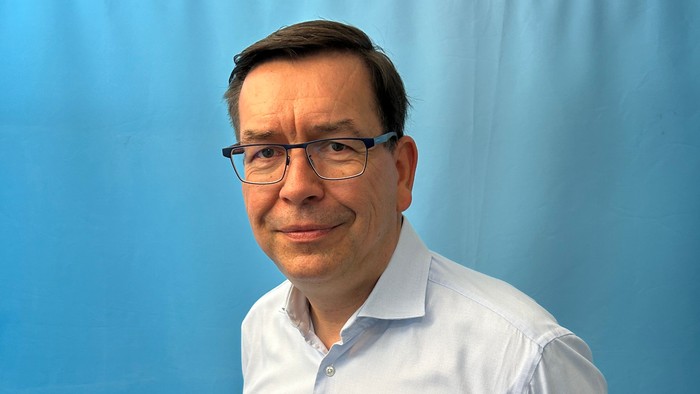 Mann mit Brille und weißem Hemd vor blauem Hintergrund.