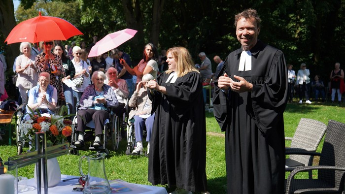 Eine Gruppe von Menschen singt im Freien, während zwei Priester in schwarzen Gewändern vorne stehen