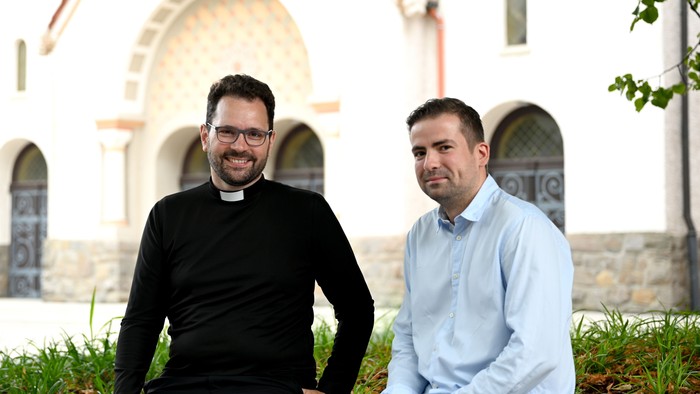 Zwei Männer sitzen vor einer Kirche und lächeln.