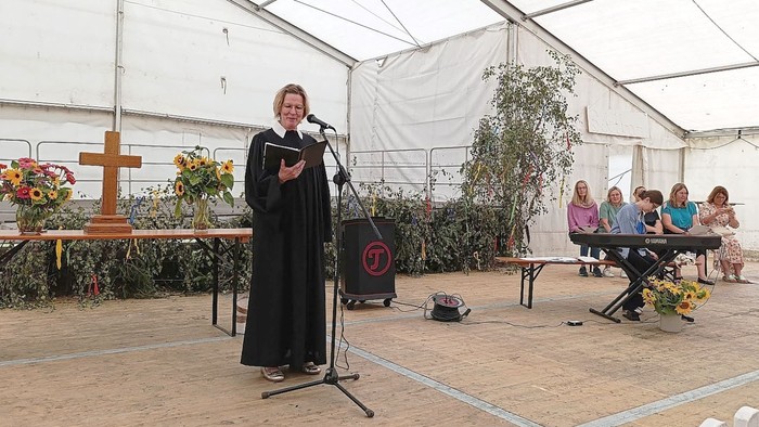 Eine Frau in einem schwarzen Gewand spricht in ein Mikrofon unter einem Zelt, während hinter ihr eine Gruppe von Menschen an einem Tisch sitzt.