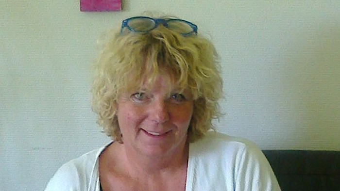 En kvinde med blond hår og briller sidder på en mørk sofa.