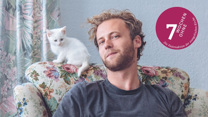 Aktionsmotiv zur Fastenaktion 7 Wochen ohne 2026: Ein Mann auf einem geblümten Sessel mit einer weißen Katze auf der Lehne