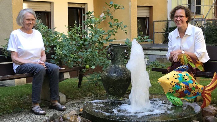 Zwei Frauen sitzen auf Bänken neben einem Springbrunnen im Innenhof eines Gebäudes.