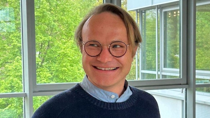 Mann in blauer Strickjacke und Jeans lächelt in einem Raum mit großen Fenstern.