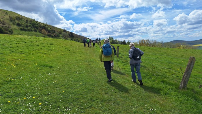 Zwei Personen wandern über eine grüne Wiese mit Wildblumen unter einem teilweise bewölkten Himmel.