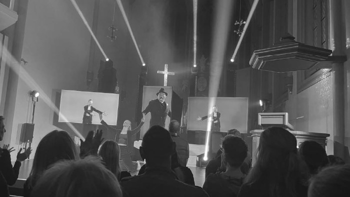 Gottesdienst mit Prediger auf der Kanzel, besucht von der Gemeinde, beleuchtet von Scheinwerfern.