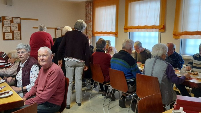 Gruppe älterer Menschen, die sich in einem Speisesaal versammelt hat – einige sitzen an Tischen mit Essen, andere stehen und unterhalten sich.