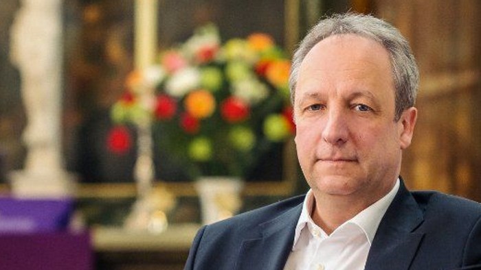 Ein Mann im Anzug sitzt nachdenklich in einer Kirche, die mit Blumen und religiösen Dekorationen geschmückt ist.