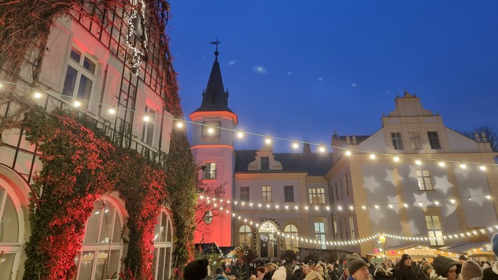 In der Abenddämmerung versammeln sich Menschenmengen auf einem Weihnachtsmarkt im Freien vor geschmückten Gebäuden mit Lichterketten.