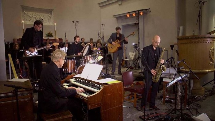En gruppe musiker spiller i et rum med forskellige instrumenter som piano, guitar og saxofon.
