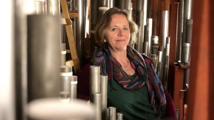 Eine Frau steht lächelnd zwischen großen industriellen Gärbottichen in einer Brauereiumgebung.