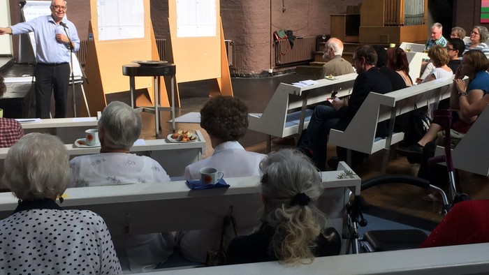 Ein Redner hält vor einem aufmerksamen Publikum einen Vortrag, das in einem kirchenähnlichen Hörsaal sitzt, während auf den Tischen Snacks bereitstehen.