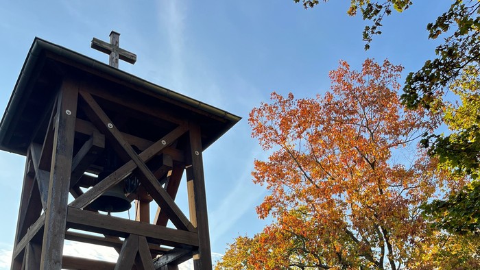 Holzkreuz auf Turm neben gelbem Haus und Herbstbaum