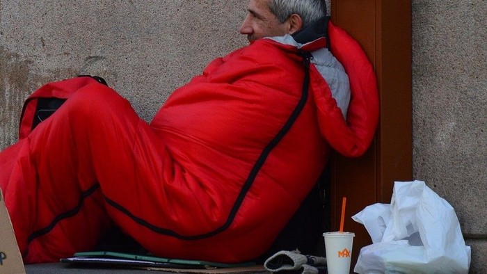 Eine Person in einem roten Schlafsack lehnt an einer Tür, mit einem Getränk und einer Tasche in der Nähe.