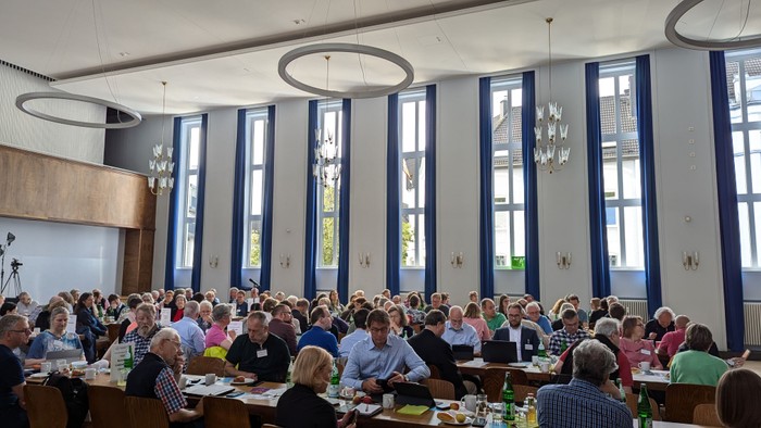 Großer Saal mit vielen Menschen an Tischen, große Fenster, Deckenleuchten