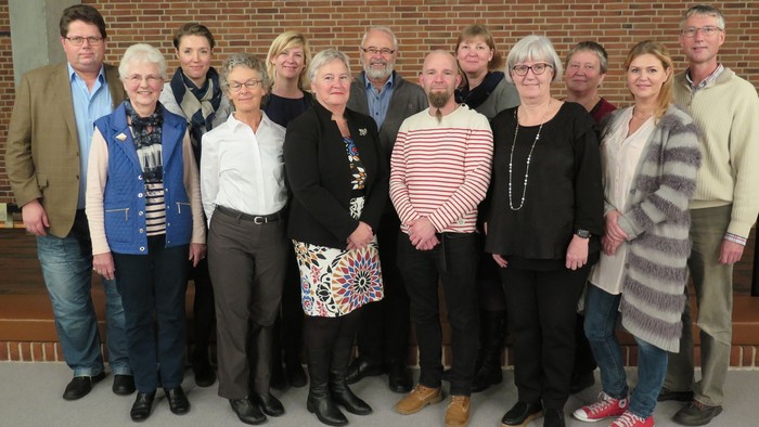 En gruppe af 14 personer står sammen til et fællesskud i et rum med mur af røde mursten.