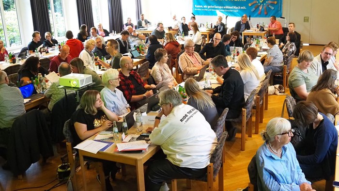 Eine Gruppe von Menschen sitzt an Tischen und arbeitet in einem Raum mit einem großen Banner im Hintergrund.