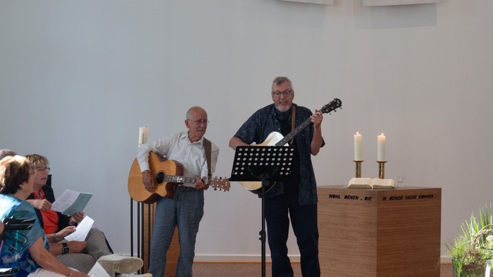 Ein Mann spielt Gitarre, während zwei andere Männer singen, eine Frau sitzt und liest.