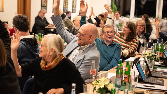 Eine Gruppe von Menschen sitzt an einem Tisch und hebt die Hände, offenbar bei einer Feier oder einem Treffen.