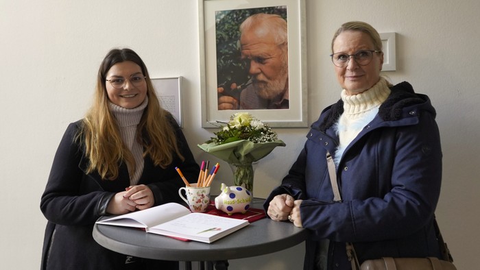 Zwei Frauen stehen an einem Tisch mit Blumen und Büchern, darüber ein Porträt.