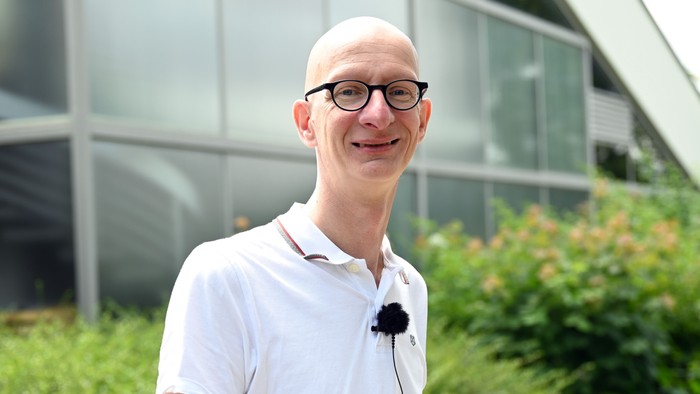 Mann in weißem Hemd vor modernem Gebäude mit Glasfront und Grünpflanzen.