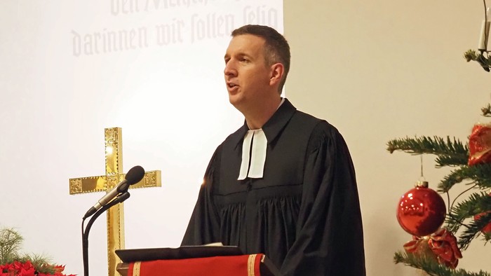 Ein Priester predigt an einem Pult mit christlichem Symbol vor einem Weihnachtsbaum.