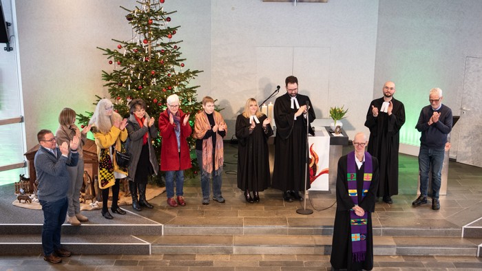 Eine Gruppe von Menschen steht vor einem Weihnachtsbaum in einem Raum, einige in Roben, andere in festlicher Kleidung.