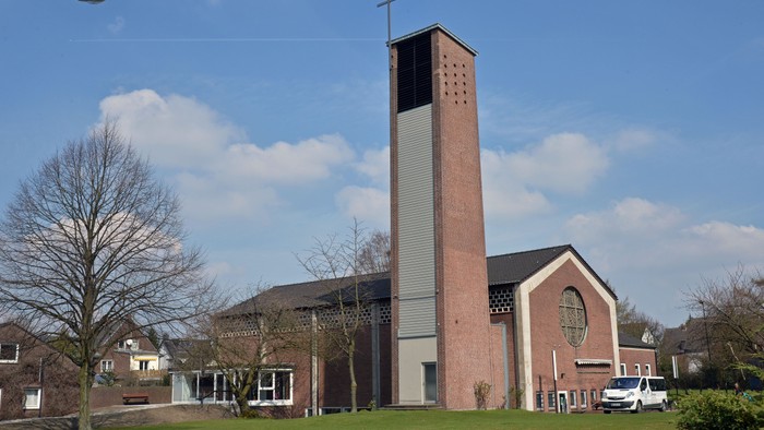 Kirchengebäude mit hohem Turm und Kreuz auf dem Dach, umgeben von Bäumen und Häusern