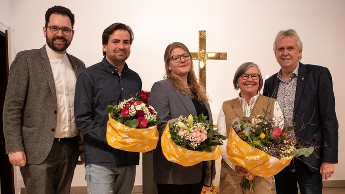 Eine Gruppe von fünf Personen steht vor einem Kreuz und hält Blumensträuße.