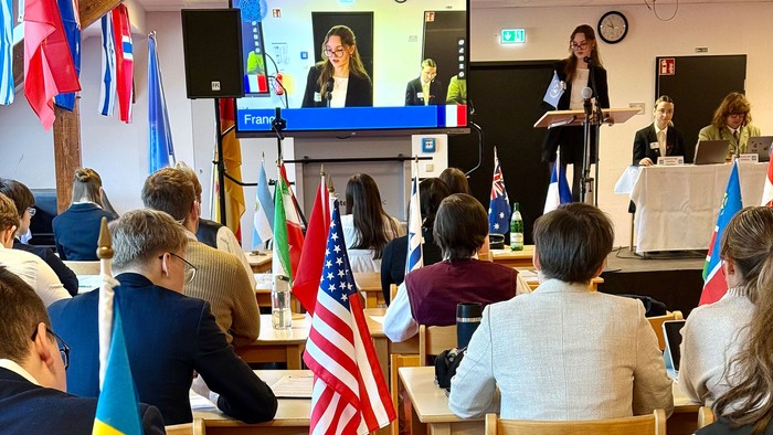 Eine Gruppe von Menschen sitzt in einem Raum mit verschiedenen Flaggen und schaut einem Vortrag auf einem Bildschirm zu.