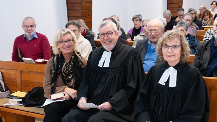 Drei Personen in Kirchenbänken, zwei in schwarzen Roben mit Perücken