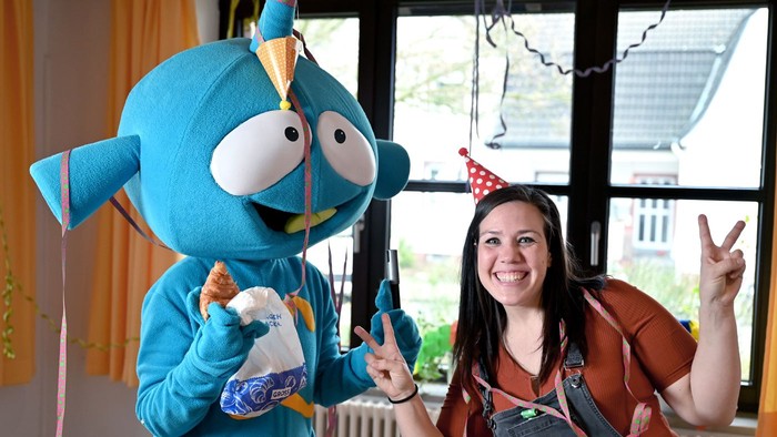 Eine Frau feiert mit einem blauen Maskottchen Geburtstag, beide tragen Partyhüte.