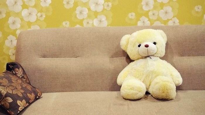 Ein gelber Teddybär sitzt auf einem beigefarbenen Sofa vor einer gelben Blumenwand.