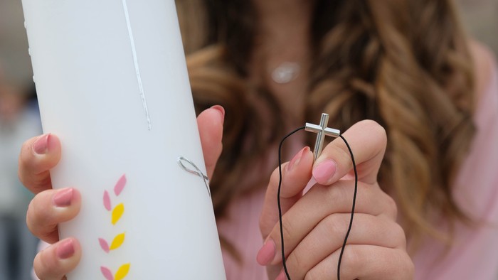 Eine Person, die eine weiße Kerze mit rosa und gelben Mustern hält und eine Kreuzkette trägt.