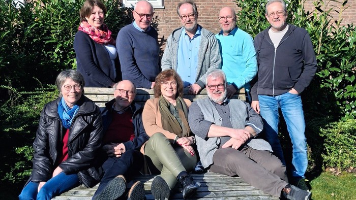 Ein Gruppe von neun Personen posiert vor einem Haus, sieben stehen und zwei sitzen auf einer Bank