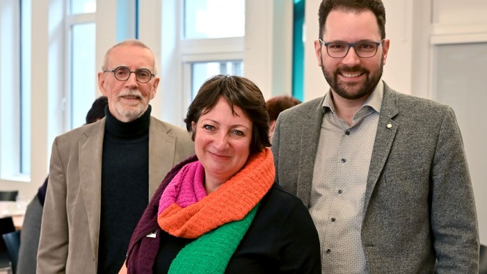 Drei Personen stehen in einem Raum mit großen Fenstern.