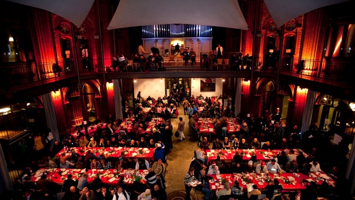 Großer Saal mit vielen Tischen und Menschen, rote Vorhänge, Bühne im Hintergrund