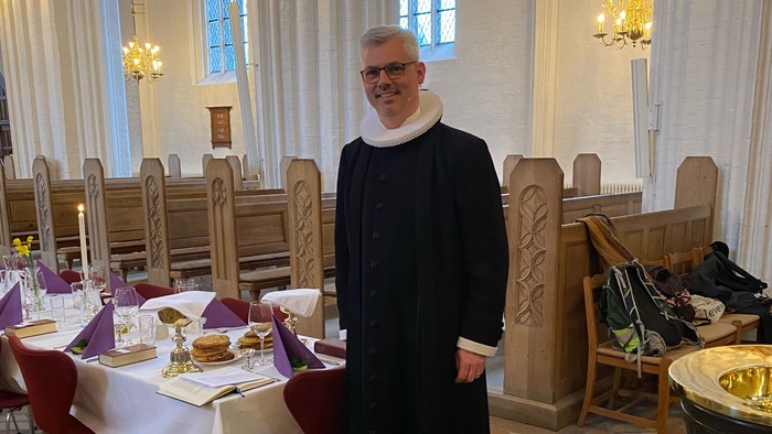 Mand i svart kapittel i kirke med bord og stoler.